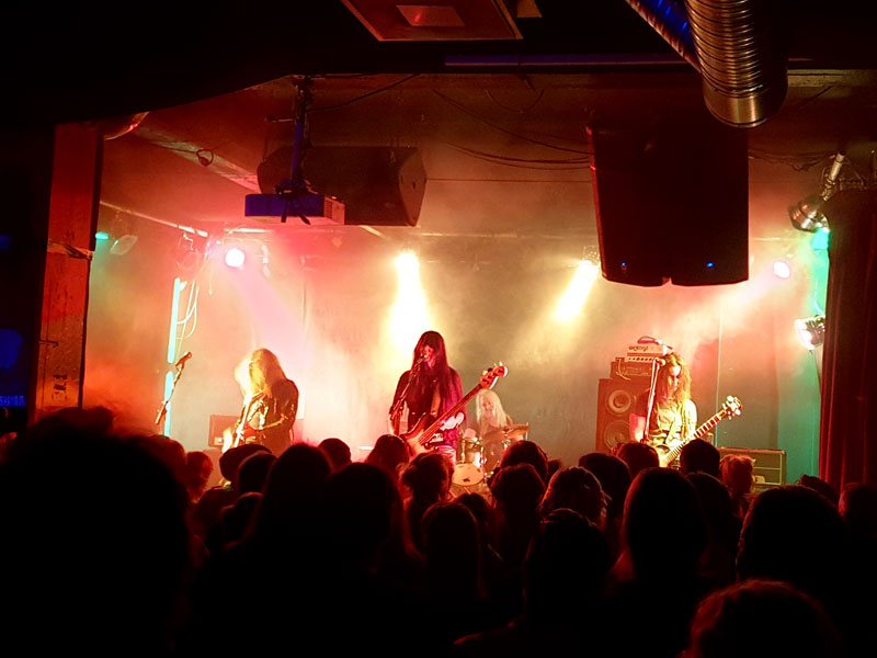 Ett band spelar på scenen. Tre personer har gitarr och bas, en trummis bakom.