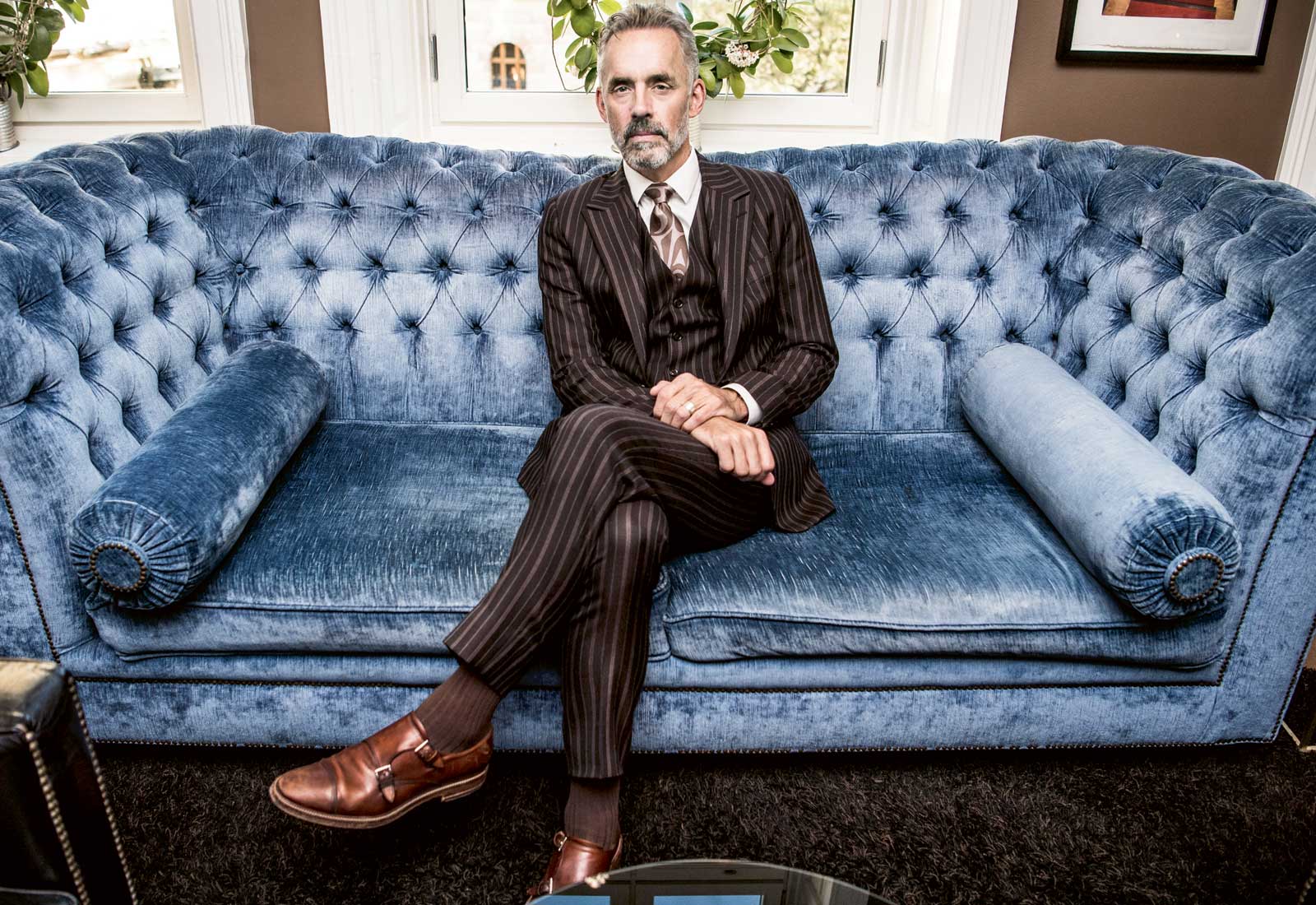 Jordan B. Peterson, i klädd en brun kostym med kritstrecksränder, i en stor blå soffa.
